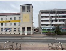 Straßenansicht eines Gebäudekomplexes mit einer Filiale der Deutschen Post auf der linken Seite, erkennbar an der gelben Beschilderung und der Sonnenuhr auf der Fassade. Rechts daneben ein mehrstöckiges Wohnhaus mit Balkonen und im Erdgeschoss Geschäfte, darunter ein „Welcome Centre“.