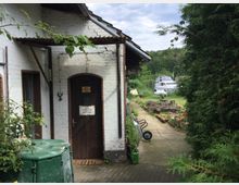 Hof mit einem weißen Ziegelgebäude, dessen Eingangstür aus Holz besteht. Im Vordergrund steht eine grüne Mülltonne, im Hintergrund sind ein Gehweg, gestapeltes Baumaterial und ein Boot auf einer Wiese zu sehen.