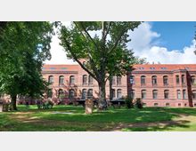 Backsteinbau mit roten Dachziegeln, großen Bogenfenstern und einem großzügigen grünen Außenbereich mit Rasenflächen und Bäumen; im Vordergrund steht ein großer Baum, Gartenmöbel sind umgeben von Schatten spendenden Bäumen aufgestellt.