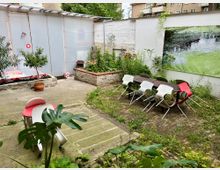 Ein kleiner Innenhofgarten mit einer überdachten Wand aus lichtdurchlässigem Material, einer Steinmauer, Blumenbeeten und einem Sitzbereich mit einem langen Tisch und mehreren Stühlen. Die Umgebung ist teilweise bewachsen, und ein großes Wandbild mit einer Naturdarstellung schmückt die weiße Rückwand.