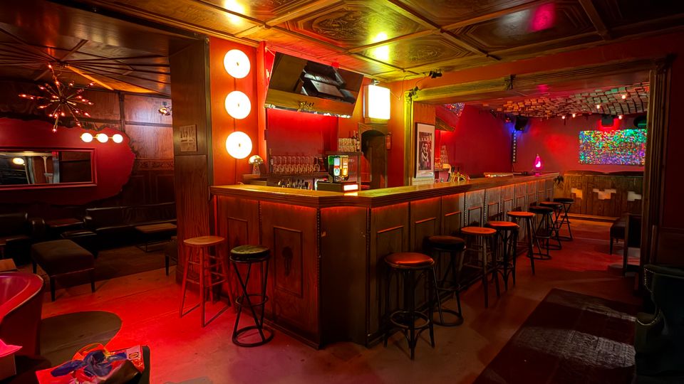 Innenraum einer Bar mit langer Holztheke und hohen Barhockern, rot-gelb beleuchtet mit dekorativen Wandlampen und einem Fernseher über dem Tresen. Im Hintergrund sind Lounge-Sitzbereiche und eine gemusterte, kassettierte Decke mit Ornamentelementen zu sehen.