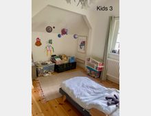Kinderzimmer mit Holzboden, einem niedrigen Bett mit weißer Bettdecke und einem ausgestopften Spielzeug darauf. An der Wand hängen bunte Bastelarbeiten, ein Modell des Sonnensystems schwebt von der Decke, und es gibt Regale mit Büchern, Spielsachen und eine Puppenhausnachbildung.
