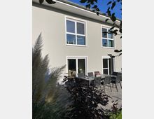 Zweistöckiges modernes Wohngebäude mit weißer Fassade und großen Fenstern. Im Vordergrund befindet sich eine Terrasse mit einem Esstisch, mehreren Stühlen sowie Pflanzen und Ziergras.