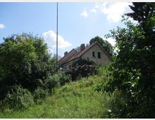 Ein einfaches Haus mit rotem Satteldach und verputzter Fassade steht auf einer leicht überwachsenen, grasbewachsenen Fläche, umgeben von dichtem Gebüsch und Bäumen. Im Vordergrund ist ein großer Stapel Holz sichtbar, die Szene ist bei sonnigem Wetter aufgenommen.