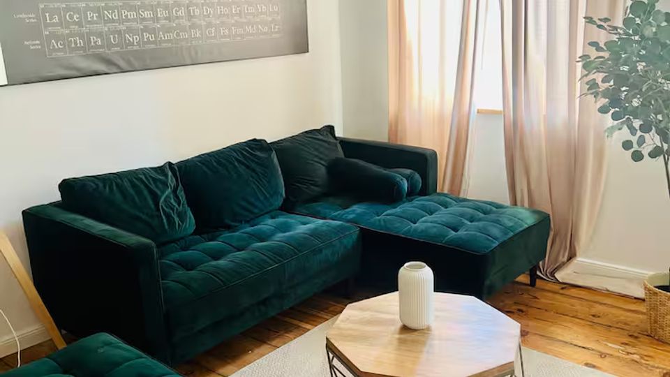 Wohnzimmer mit dunkelgrünem, gepolstertem Ecksofa und passendem Hocker auf hellem Teppich, davor ein achteckiger Couchtisch mit Holzplatte und Metallgestell. An der Wand hängt ein großes Periodensystem-Poster; daneben ein Fenster mit bodenlangen, beigen Vorhängen und Holzboden sowie eine Zimmerpflanze in der Ecke.