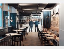 Ein modernes Café mit einer Mischung aus Sitzgelegenheiten an kleinen Tischen und einer gepolsterten Sitzbank an der Wand; die Rückwand ist mit einer großen Wandgrafik verziert, und die Bar im Hintergrund ist mit Kaffeemaschinen und Regalen ausgestattet. Schwarze Fliesen und dezente Hängeleuchten prägen die stilvolle Atmosphäre.