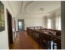 Ein heller Flur in einem historischen Gebäude mit einem Holzgeländer, das eine Treppe umrahmt. Die hohen Fenster lassen viel Tageslicht herein, und die weiße Stuckdecke sowie Holzböden und rote Teppiche unterstreichen den klassischen Stil.