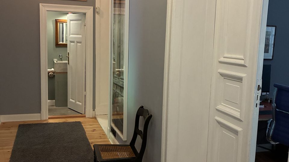 Langer Flur in einer Wohnung mit grauen Wänden, Holzdielenboden und dunklem Läuferteppich, beleuchtet von hängenden Glühbirnen. Weiße Zimmertüren und Türrahmen, gerahmte Bilder an der Wand sowie ein Stuhl neben einer Vitrine; am Ende ist ein kleines Bad sichtbar.