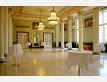 Ein prächtiger, klassischer Festsaal mit hohen, verzierten Säulen, großen Kristalllüstern und bodentiefen Fenstern. Der Raum ist mit kleinen, weiß gedeckten Stehtischen ausgestattet und hat eine hellbeige Marmorfassade.