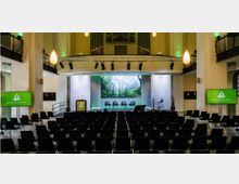 Veranstaltungssaal mit Reihen schwarzer Stühle, einer Bühne mit Podium, mehreren Stühlen und einem großen Bildschirm mit einem Naturmotiv im Hintergrund. Der Raum hat hohe Decken, Hängebeleuchtung, eine Empore mit Geländern und seitliche Bildschirme mit dem Logo „Besondere Orte“.