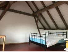 Gemütlicher Dachbodenraum mit sichtbaren Holzbalken, weiß gestrichener Decke und Wänden. Im Raum steht ein schwarzes Metallbett mit weißer Bettwäsche und eine schlichte Holztischplatte, der Boden ist rot.