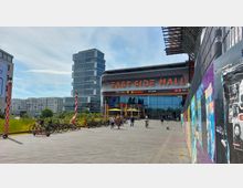Einkaufszentrum mit der Beschriftung „East Side Mall“ auf der Fassade, flankiert von modernen Bürogebäuden. Im Vordergrund befinden sich Fahrräder, Passanten und bunte Plakatwände auf der rechten Seite.