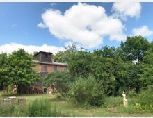 Backsteingebäude mit einem Dachaufbau aus dunklem Holz, umgeben von dichtem, grünem Baumbestand und einem Garten mit wilder Vegetation; im Vordergrund eine Skulptur einer sitzenden Figur und einige Steinblöcke auf einer Wiese. Der Himmel ist klar mit vereinzelten weißen Wolken.