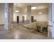 Treppe in einem öffentlichen Gebäude mit Wänden und Säulen aus Naturstein. Der Bereich ist hell beleuchtet durch moderne, runde Hängelampen, und mehrere Türen führen zu angrenzenden Räumen.