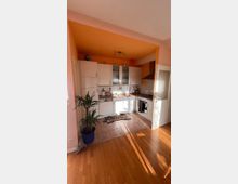 Eine kleine, offene Küche mit weißen Schränken, brauner Arbeitsplatte und Fliesenspiegel. Der Raum hat einen orangefarbenen Deckenabschnitt, Holzboden im angrenzenden Bereich, und eine grüne Zimmerpflanze steht in einem blauen Topf am Übergang zur Küche.