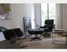 Wohnzimmer mit minimalistischer Einrichtung, darunter ein schwarzer Schaukelstuhl, ein schwarzer Ledersessel mit Fußhocker und ein kleiner Beistelltisch. Im Hintergrund befindet sich ein großes Fenster mit Blick auf grüne Pflanzen.