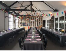 Ein industriell gestalteter Veranstaltungssaal mit hohen Decken und sichtbaren Metallträgern. Lange, mit dunkelgrauen Tischdecken bedeckte Tische sind festlich gedeckt, und bunte Wimpelketten schmücken den Raum. Große Fenster und Holzelemente an der Stirnwand prägen die Atmosphäre.