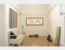 Heller Flur mit Parkettboden, einer weißen Holzbank mit abgelegten Gegenständen und Schuhen darunter, einem großen rechteckigen Spiegel mit goldfarbenem Rahmen an der Wand sowie einem abstrakten, bunt gestreiften Gemälde. Entlang der Wände stehen weitere Schuhe und ein gerahmtes Bild auf dem Boden.