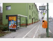 Bushaltestelle mit einer überdachten Sitzbank und Reklame für einen Radiosender („104.6 RTL“) im Vordergrund. Im Hintergrund verläuft eine Straße mit einem Geh- und Radweg entlang eines langen, grün gestrichenen Mehrfamilienhauses. Ein orangener Mülleimer und ein Haltestellenschild sind ebenfalls sichtbar.