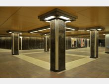 Unterirdischer U-Bahn-Bereich mit rechteckigen Pfeilern, die beleuchtete Decken tragen. Die Wände sind mit dunklen Fliesen verkleidet, und an einigen Stellen sind Werbeplakate angebracht.