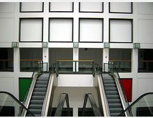 Innenraum eines modernen Gebäudes mit symmetrischem Design, zwei Rolltreppen, die nach oben führen, und weißen Wänden mit quadratischen Fensterelementen. Im Hintergrund sind bunte Türen (eine grün, eine rot) und Glasgeländer sichtbar.