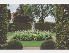 Geometrisch angelegter Garten mit einer zentralen runden Blumenrabatte, in deren Mitte ein kleiner Springbrunnen steht. Umgeben von gepflegten Buchsbaumhecken und einem Kiesweg sowie hochgewachsenen Bäumen im Hintergrund.