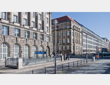 Straßenszene mit historischen Gebäuden aus hellem Stein, hohen Fenstern und klassischer Architektur. Im Vordergrund eine U-Bahn-Eingangstafel mit der Aufschrift „Mohrenstraße“, daneben eine gepflasterte Gehwegfläche und vereinzelte Passanten.