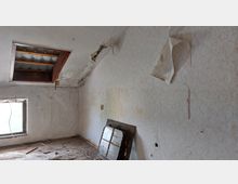 Ein heruntergekommener Dachraum mit beschädigter Wand- und Deckenverkleidung, sichtbarer Feuchtigkeit und abgeblätterter Tapete. Ein defektes Dachfenster und ein kleines bodennahes Fenster lassen Tageslicht herein, während am Boden Teile von Bauschutt und Kabeln liegen.