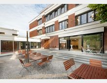 Moderne Gebäude mit Backsteinfassade und großen Fenstern, umgeben von einem gepflasterten Außenbereich mit Holz-Tischen und -Stühlen. Eine Treppe verbindet den Hof mit den bodentiefen Fenstern und Eingängen des Gebäudes, das durch weißen Putz akzentuiert ist.