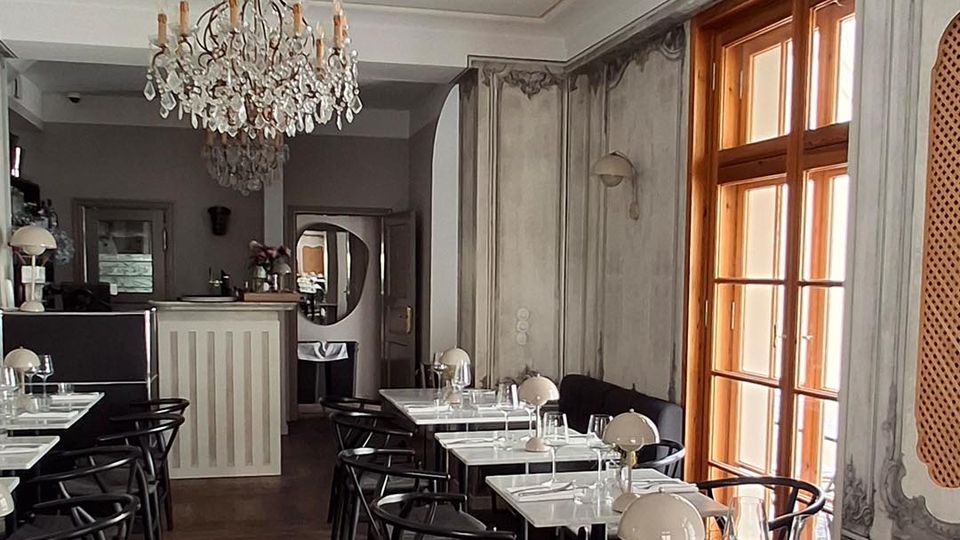 Eleganter Café- oder Restaurantinnenraum mit Kronleuchtern, dunklen Holzstühlen und weiß gedeckten Tischen auf Fischgrätparkett. Rechts sorgen hohe Holzfenster und klassisch dekorierte Wandpaneele in Grautönen für Tageslicht und einen historischen Ambientecharakter.
