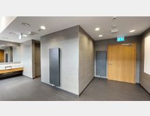 Modernes Badezimmer mit grauen Fliesen, einem Waschtisch mit Spiegel und einem hölzernen Unterschrank. Im Raum befinden sich eine helle Holztür mit Fluchtweg-Schild darüber und flache, schwarze Heizkörper an den Wänden.