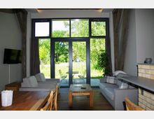 Wohnzimmer mit großen, bodentiefen Fenstern, die auf eine Terrasse mit Gartenmöbeln und Blick auf eine grüne Wiese führen. Der Raum ist mit Holzfußboden, einer grauen Sofagarnitur, Holztisch, Esstisch mit Stühlen und einer gemauerten Küchenbar ausgestattet.