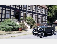 Fachwerkhaus mit hellem Putz und dunkler Holzverkleidung, vor dem Eingang eine Treppe mit Blumen und Sträuchern. Im Vordergrund steht ein dunkelgrünes Oldtimer-Auto auf einer Straße.