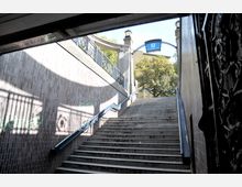 Treppenaufgang einer U-Bahn-Station mit geländerlosen Stufen, flankiert von Backsteinwänden. Oben befindet sich das Schild "U Heidelberger Platz", daneben Bäume und ein mit Geländern verzierter Steinbogen.