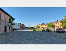 Ein offener Platz mit Kopfsteinpflaster, umgeben von historischen Backsteingebäuden, teils mit Pflanzen bewachsen. Im Hintergrund sind parkende Autos, Grünflächen mit Bäumen und vereinzelte Laternen zu sehen, unter klarem Himmel.