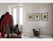 Ein heller Eingangsbereich mit einer weiß gerahmten Fensterfront. Links steht eine Garderobe mit Kleidung, Taschen und Hüten, rechts hängen drei gelb-schwarze Kunstwerke in braunen Rahmen an der Wand, und darunter befindet sich eine weiße Kommode mit einem geflochtenen Korb.