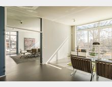 Helles, modernes Büro mit großen Fensterfronten und Blick auf Bäume. Im Vordergrund stehen zwei Sessel an einem Glastisch, im Hintergrund befindet sich ein Konferenzraum mit mehreren Bürostühlen und einem großen Gemälde an der Wand.