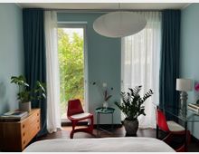 Modernes Schlafzimmer mit hellblauen Wänden, zwei bodentiefen Fenstern mit weißen Gardinen und dunklen Vorhängen sowie Blick ins Grüne. Im Raum stehen rote Stühle, ein Glastisch mit Lampe, mehrere Zimmerpflanzen, eine Holzkommode und eine große runde Papier-Deckenleuchte.