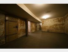 Ein spärlich beleuchteter Kellerraum mit abgenutzten Wänden und mehreren hölzernen Zellentüren auf der linken Seite. Der Boden ist aus Beton, und das Licht einer Deckenlampe erhellt den Raum schwach.