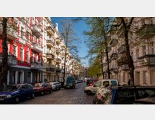 Straßenszene mit gepflastertem Straßenbelag, flankiert von mehrstöckigen Altbauten mit verzierten Balkonen in Weiß- und Rottönen. Parkende Autos säumen die Straße, während Bäume mit frischem Grün den Gehweg begrenzen.