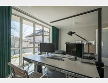 Modernes Büro mit großer Fensterfront, die den Blick auf einen Garten und ein benachbartes Gebäude freigibt. Im Raum stehen ein Schreibtisch mit zwei Monitoren, eine Schreibtischlampe und ein Drehstuhl; die Vorhänge sind grün und der Boden grau.