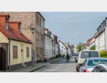 Eine ruhige Wohnstraße mit geparkten Autos auf beiden Seiten, flankiert von mehrstöckigen Wohnhäusern mit Putz- oder Ziegelfassaden und teils roten Ziegeldächern. Auf dem Gehweg sind Straßenlaternen und vereinzelte Fußgänger und Radfahrer zu sehen.