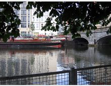 Blick auf einen Fluss mit reflektierendem Wasser, im Vordergrund Zweige eines Baumes und ein Metallzaun. Am Flussufer liegen rote Lastkähne vor einer Steinbrücke mit zwei Bögen, dahinter moderne Gebäude.