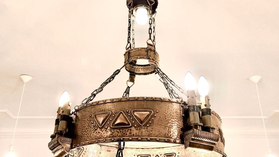 Großer, an Ketten aufgehängter Kronleuchter aus Metall im Industrie-/Steampunk-Stil mit genieteten Ringen und mehreren freiliegenden Glühbirnen. Hängt an einer hellen, weißen Decke mit weiteren kleinen Deckenleuchten im Hintergrund.