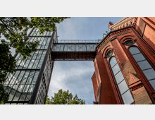 Ansicht eines modernen Gebäudeturms aus Glas und Stahl, verbunden durch einen verglasten Übergang mit einer roten Backsteinfassade eines historischen Gebäudes mit großen Fenstern und dekorativen Elementen. Im Vordergrund sind Äste und Blätter eines Baumes zu sehen, und der Himmel ist leicht bewölkt.