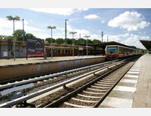 Bahnhof mit Gleisen, Bahnsteigen und einem wartenden roten S-Bahn-Zug. Im Hintergrund sind überdachte Bahnsteige, Laternen und ein Werbeplakat zu sehen, umgeben von Bäumen und klarem Himmel.