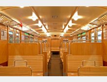 Innenraum eines historischen Zugwagons mit hölzernen Sitzbänken, Holzverkleidungen an den Wänden und drahtgitterartigen Ablagen über den Sitzen. Die Decke ist hell gestaltet, und seitlich sind Fenster sowie Hinweisschilder angebracht.