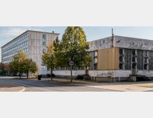 Ein urbanes Straßeneck mit einem sanierungsbedürftigen, rechteckigen Gebäude mit großen Glasbausteinfenstern und einem angrenzenden mehrstöckigen Flachbau mit weißen Fassaden. Im Vordergrund sind Bäume, ein Gehweg und ein Straßenschild zu sehen.