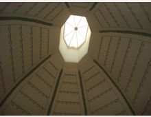 Blick auf eine oktogonale Kuppeldecke mit einem zentralen, achteckigen Oberlicht, das Tageslicht einlässt. Die Decke ist mit dekorativen Vertiefungen und grünen, pflanzenartigen Verzierungen gestaltet.