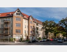 Mehrstöckiges Wohngebäude im Stil der frühen 20. Jahrhunderts mit hellbrauner und beige verputzter Fassade, zahlreichen Fenstern und kleinen Balkonen, daneben auf der Straße parkende Autos und Bäume am Straßenrand.